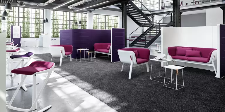 Das Foto zeigt eine einladende, moderne Büroumgebung mit dunklem Treppenaufstieg mit Glasgeländer im Hintergrund und industriellen Fenstern. Auf einem großen, anthrazitfarbenen Teppich stehen Workbays, die dank ihrer hohen Rücklehne mehrere Sitzmöglichkeiten voneinander trennen und nicht nur einen Raum für Diskretion schaffen, sondern auch die Akustik optimieren. Die Sofas und Sessel sind gepolstert und in verschiedenen Farben erhältlich – fragen Sie Wünsche GmbH für Polster in Ihren Firmenfarben!