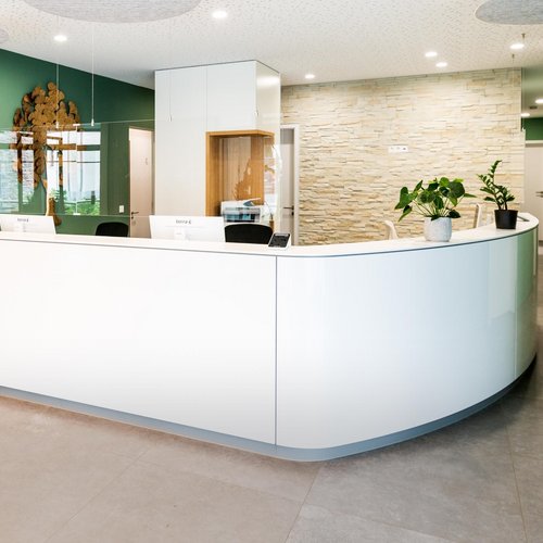 Das Foto zeigt die einladende Rezeption einer Arztpraxis. Die helle Empfangstheke in weißer Optik wurde nach Maß in der Tischlerei Wünsche GmbH gefertigt und endet nach einer länglichen Seite in einer abgerundeten Form. Pflanzen stehen auf der glatten Oberfläche des Empfangs und transparentes Acrylglas schwebt zur Sicherheit von Patienten und Mitarbeitern über der Theke. An der Decke regulieren runde Akustikpaneele den Raumklang des Foyers. In einer Säule hinter dem Empfang wurde in einer holzverkleideten Nische der Kopierer platziert.