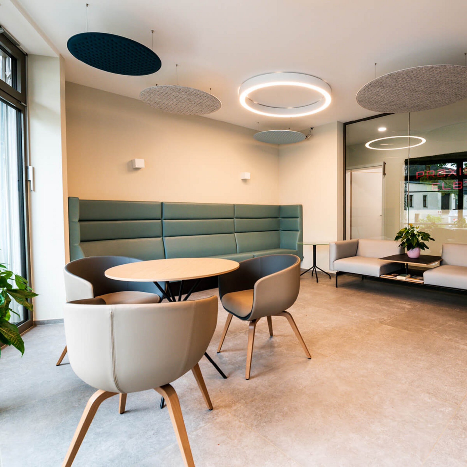 Foto eines freundlichen, hellen Wartezimmers einer Arztpraxis mit Pflanzen und bequemen, modernen Möbeln nach Maß. Ein deckenhohes Fenster lässt großzügiges Licht in den Raum. Von der Decke hängen Akustikpaneele, um den Raumklang zu optimieren.