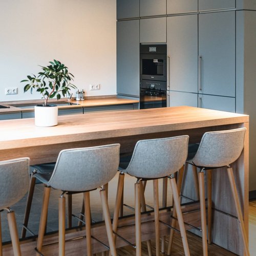 Das Foto zeigt eine moderne, minimalistische Büroküche mit einer eleganten Holztheke und gepolsterten Barhockern, die ideal für Personalräume und Kantinen geeignet ist. Die Küche verfügt über integrierte Schränke in mattem Grau, die viel Stauraum bieten, sowie moderne Küchengeräte, die in die Schrankwand eingelassen sind. Atmosphäre. Ideal für Unternehmen, die Wert auf ein stilvolles und funktionales Design in ihren Personalräumen legen.