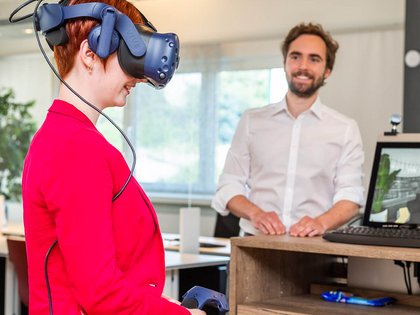 Das Foto zeigt einen freundlichen Berater der Wünsche GmbH im weißen Hemd, der aufmerksam eine Kundin im Blazer beobachtet, die eine VR-Brille trägt und so eine virtuelle Begehung ihrer geplanten Räumlichkeiten macht. 