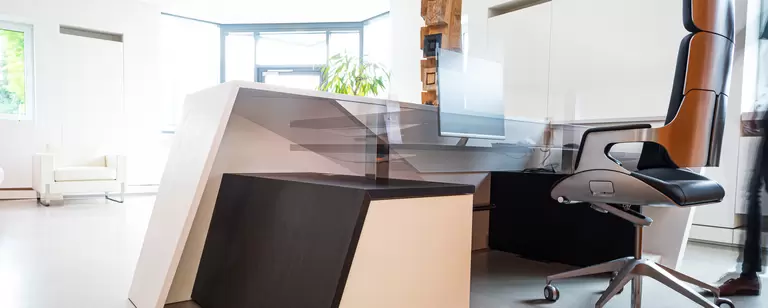 Das Bild zeigt den modernen, minimalistischen Empfangsbereich der Wünsche GmbH. Im Vordergrund befindet sich ein stilvoller Empfangstresen mit klaren, geometrischen Linien in Weiß und dunklem Holz. Hinter dem Tresen steht ein ergonomischer Bürostuhl mit Metall- und Lederdetails. Die Umgebung ist hell gestaltet, mit viel natürlichem Licht, das durch große Fenster einfällt. Eine weiße Sitzgruppe und eine grüne Pflanze im Hintergrund sorgen für eine einladende Atmosphäre. 