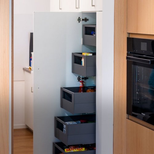 Aus einer Küche mit Holzmöbeln nach Maß wird in den angrenzenden Hauswirtschaftsraum fotografiert. Der von Wünsche GmbH geplante Stauraum wurde in der hauseigenen Tischlerei umgesetzt und beim Kunden fachgerecht montiert. Hinter weißen Schränken verbirgt sich viel Platz für Speisekammer und hauswirtschaftliche Geräte. Ein lautloses Schubladensystem garantiert eine aufgeräumte und platzsparende Aufbewahrung.